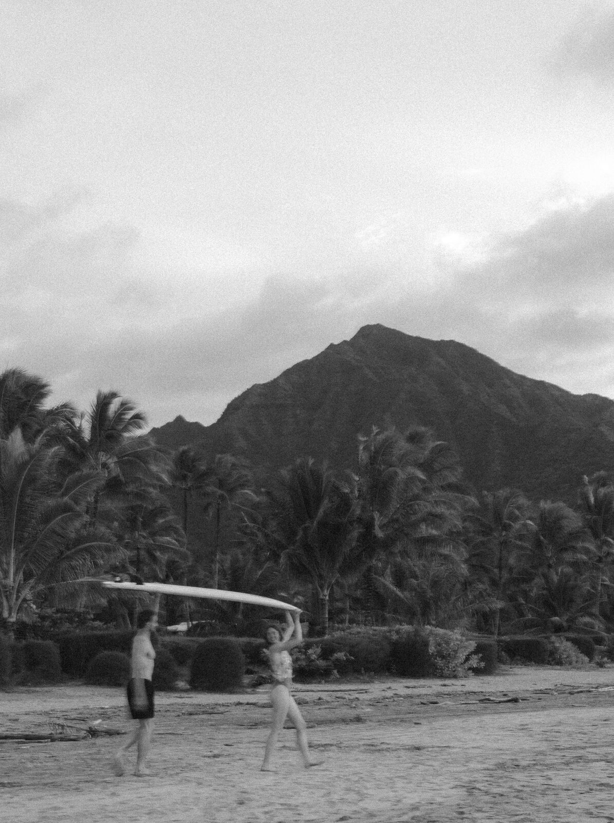 Carley & Tommy — a surfer crossing the beach below a green mountain, black and white