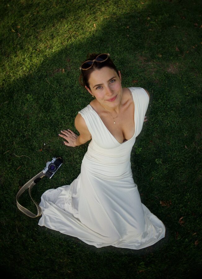 Delani McKee in a white dress on the grass beside her vintage Canon film camera