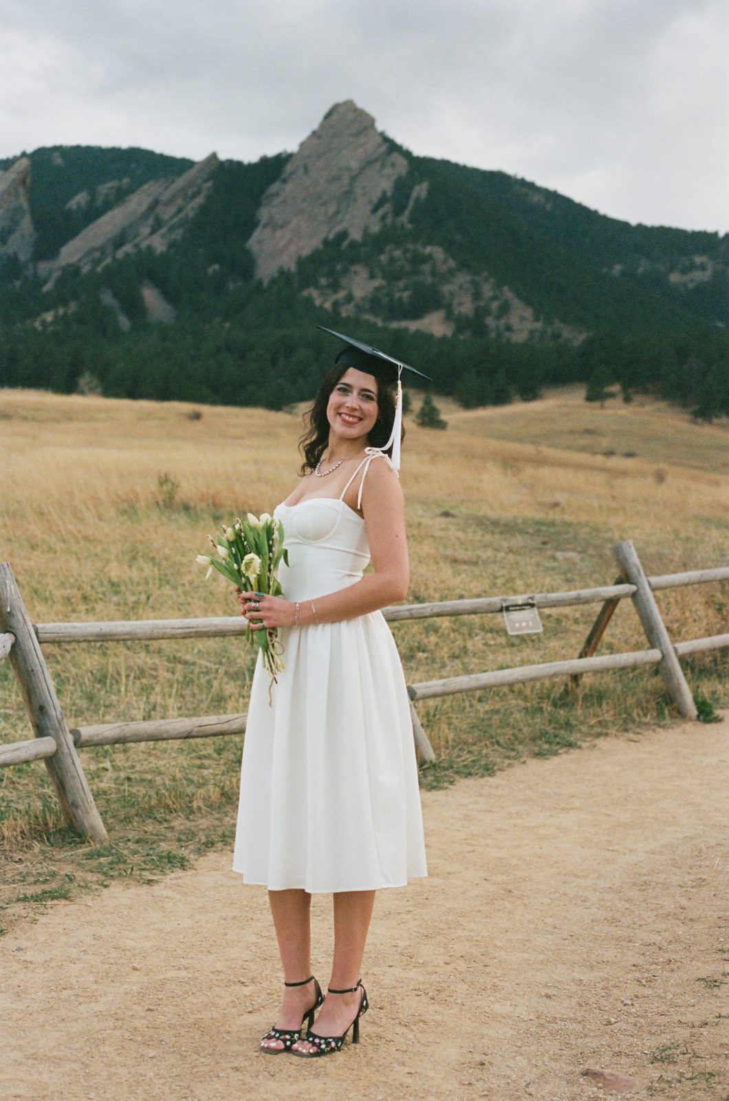 A graduate in a white dress and cap holding flowers beneath the Flatirons