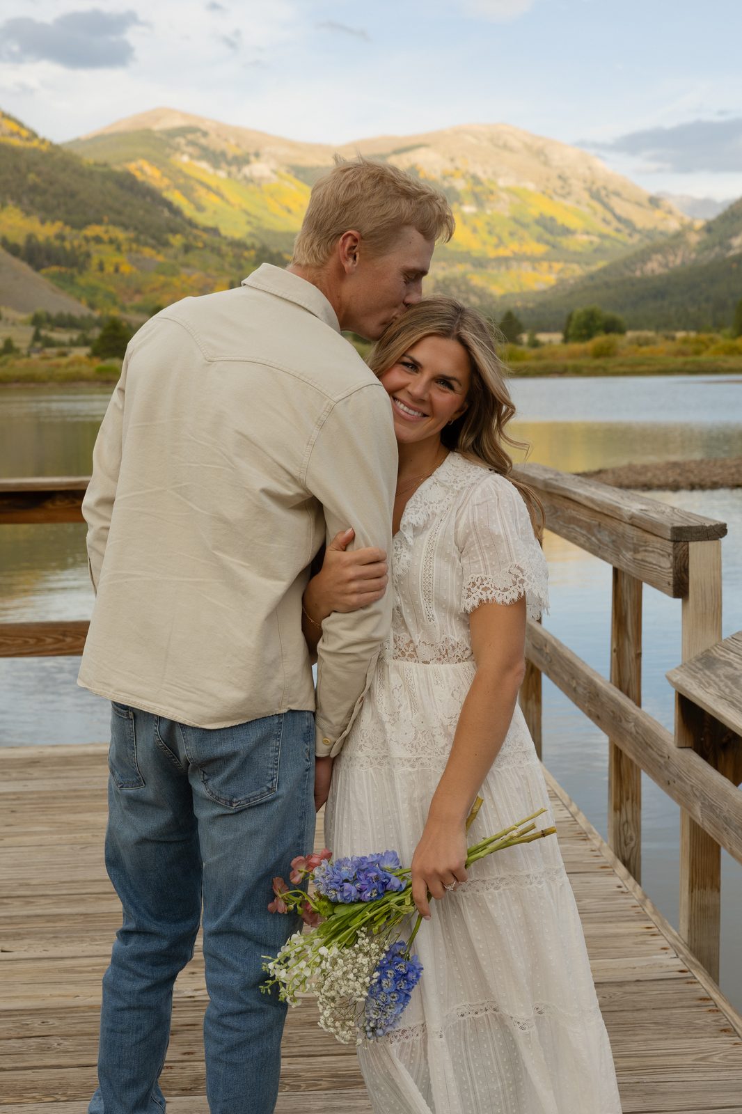 Couple embracing on a lakeside boardwalk beneath golden mountains