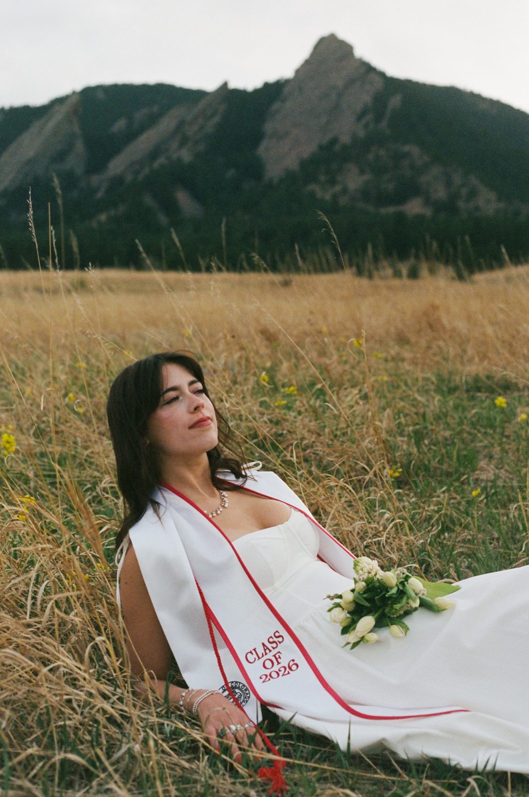 A graduate with a sash and flowers lying in a meadow below the Flatirons